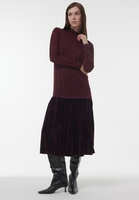 Frau in einem langärmeligen burgunderfarbenen Oberteil, einem samtigen dunkelvioletten Midikleid und schwarzen Stiefeletten mit Absatz, die mit einer Hand in der Nähe ihres Gesichts posiert.