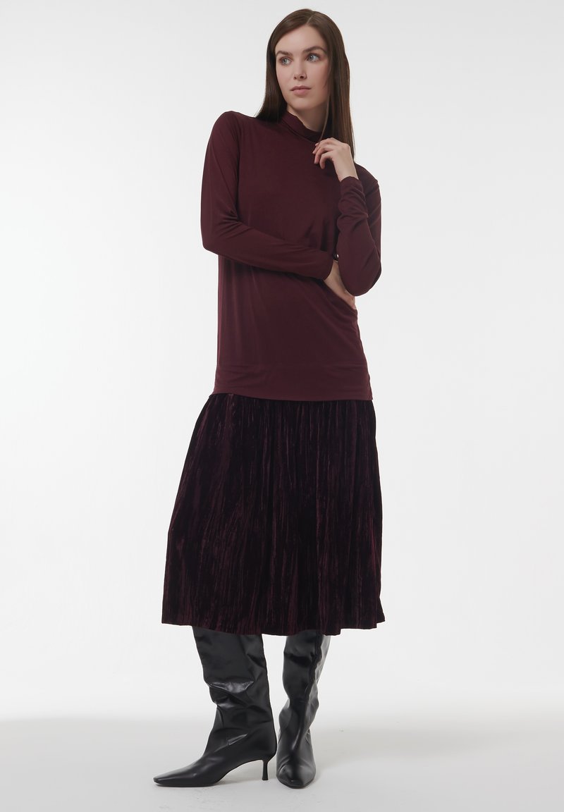 Frau in einem langärmeligen burgunderfarbenen Oberteil, einem samtigen dunkelvioletten Midikleid und schwarzen Stiefeletten mit Absatz, die mit einer Hand in der Nähe ihres Gesichts posiert.