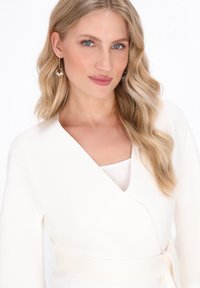 Biała bluzka z dekoltem w kształcie litery V, długimi rękawami i paskiem do związania. Modelka ma długie, faliste blond włosy i nosi okrągłe, metalowe kolczyki.