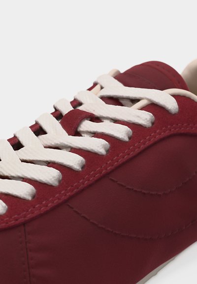 Κοντινό πλάνο ενός κόκκινου αθλητικού παπουτσιού με άσπρες κορδόνια, δείχνοντας λεπτομέρειες ραφής και υφή υφάσματος στην πλευρά.