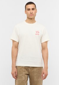 T-shirt beige clair en coton avec des manches courtes, présentant un design graphique rouge sur le côté gauche de la poitrine. Le modèle porte un pantalon texturé marron.