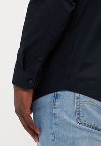 Bras droit d'une personne portant une chemise noire à manches longues et un jean en denim bleu clair, main détendue sur le côté.