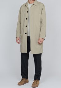 Beige, knälång kappa med rak skärning, som har svarta knappar, en spetsig krage och långa ärmar. Bärs över en ljus tröja och mörka byxor.