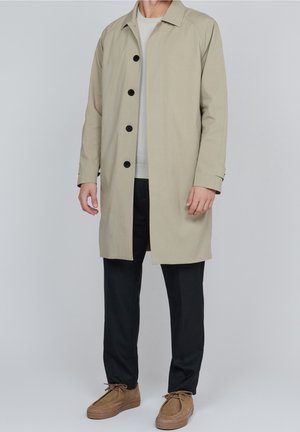 Beige, knielanger Mantel mit geradem Schnitt, schwarzen Knöpfen, spitzen Kragen und langen Ärmeln. Getragen über einem hellen Pullover und dunklen Hosen.