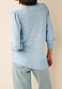 Pull en maille bleu clair avec des manches bouffantes et un ourlet côtelé, présentant un motif texturé. Assorti à un jean en denim clair.