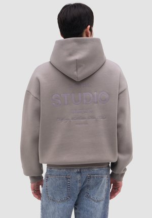 Homme portant un sweat à capuche gris avec le texte "STUDIO" au dos, associé à un jean bleu, dos tourné sur un fond uni.