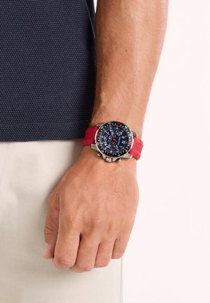 Sat s crvenim silikonskim remenom, kućištem od nehrđajućeg čelika u srebrnoj boji, okruglim brojčanikom, plavim naglascima i višestrukim kronografskim podbrojčanicima prikazan na zapešću.