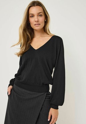 Pull noir à col en V avec des manches longues, des poignets et un ourlet côtelés. Porté avec une jupe grise à rayures fines. Texture douce, coupe décontractée.