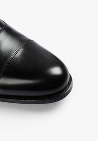 Zapato de cuero negro con acabado brillante, punta redondeada y detalle de costuras sutiles a lo largo de la parte superior. Suela plana con borde liso.