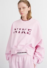 Svetloružová mikina Nike s veľkým burgundským logom; voľný strih, rebrované manžety a klasický výstrih; v kombinácii s rovnakými teplákmi.