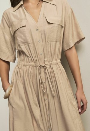 Femeie purtând o rochie bej cu nasturi, mâneci scurte, buzunare la piept, talie cu șnur și o brățară mare la încheietura mâinii stângi.