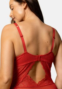 Bodysuit en dentelle rouge avec des bretelles réglables, une fermeture à crochet et des bords festonnés avec un dos ouvert. Motif texturé visible.