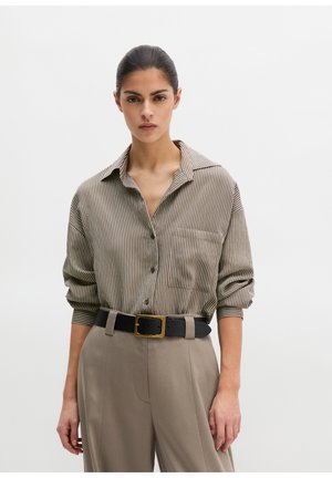 Frau mit dunkel zurückgebundenen Haaren trägt ein beiges und weiß gestreiftes Hemd mit Knöpfen, das in beige hoch taillierte Hosen mit einem schwarzen Gürtel gesteckt ist.