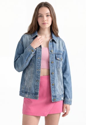 Ung kvinde iført en blå denimjakke over en pink ribbet crop top og et klart pink nederdel, stående mod en hvid baggrund.