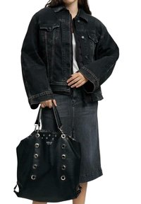 Femme portant une veste en jean noir et une jupe, tenant un sac à main Replay noir studded avec œillets argentés et doubles poignées.