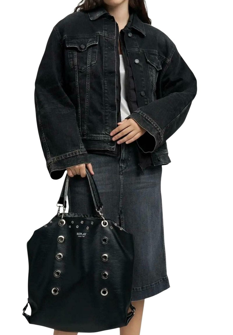 Femme portant une veste en jean noir et une jupe, tenant un sac à main Replay noir studded avec œillets argentés et doubles poignées.