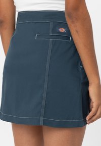 Tmavě modrá denimová sukně s bílým šitím, zadní kapsou a značkovou visačkou. Tkanina vypadá pevně a je navržena v jednoduchém rovném střihu.