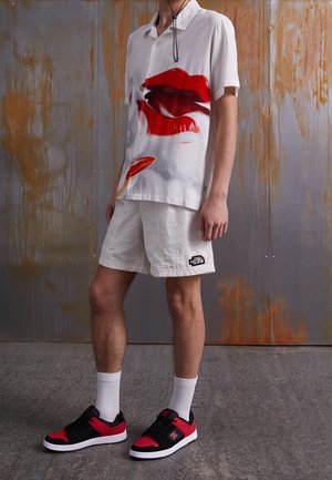 Person iført hvide shorts med The North Face-logo, en hvid skjorte med stort rødt læbeprint, hvide sokker og sort-røde sneakers.