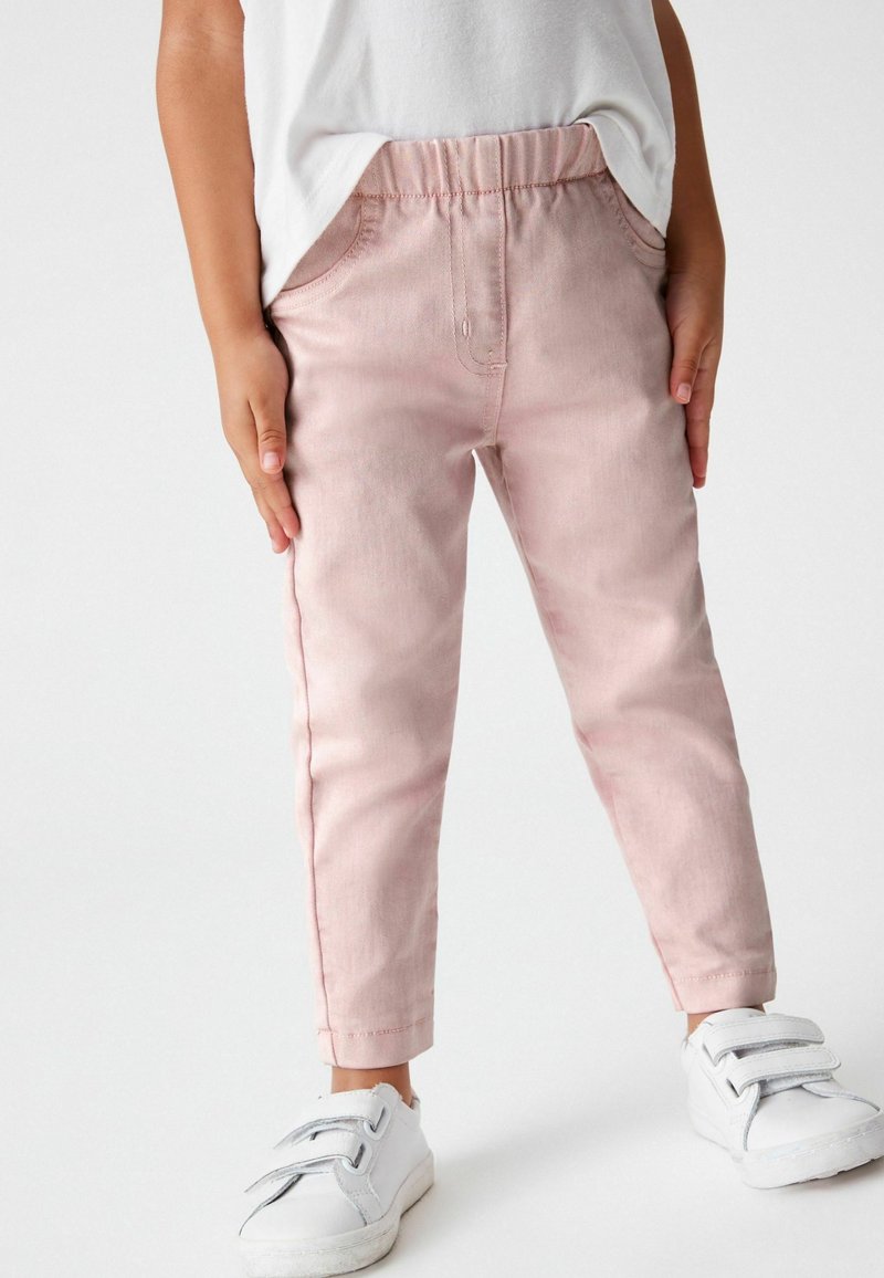 Enfant portant un pantalon à taille élastique rose clair associé à des baskets blanches à fermeture velcro et une chemise blanche.
