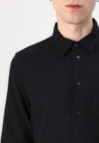Black button-up shirt with a collar, made of a textured fabric. Features a chest pocket and black buttons along the front placket.