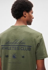 Oliivinvihreä puuvillainen T-paita, jossa on pyöreä kaula-aukko. Edessä teksti "stockholm ATHLETES CLUB" tummansinisin kirjaimin ja selässä erilaisia graafisia yksityiskohtia.