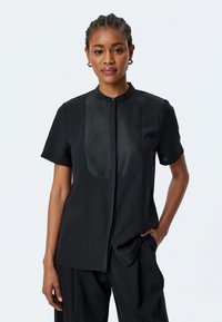 Camisa negra de manga corta con una textura suave y un panel de satén en contraste en la parte frontal. Presenta un cuello mao y cierre de botones.