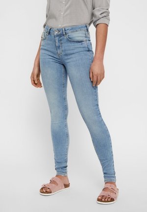 Vrouw draagt lichtblauwe strakke spijkerbroek, een lichtgrijs overhemd met opgerolde mouwen en roze sandalen met gesp.