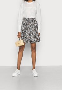 Weiße Bluse mit langen Ärmeln und strukturierten Punkten, schwarzer Blumenrock mit Rüschensaum und weiße Plateau-Sneaker. Hält eine kleine beigen Handtasche.