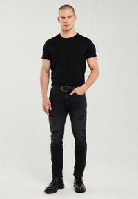 Mees seisab, kandes musta kitsast T-särki, katkendlikke musti teksapükse vöögaga ja musta pitsiga saapaid helehallil taustal.