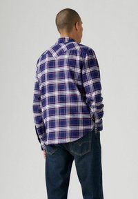 Camicia a quadri blu con accenti bianchi e rossi. Presenta maniche lunghe, colletto e orlo curvo. Indossata con jeans blu scuro.