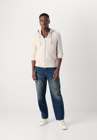 Lys beige zip-opp hettejakke med snor, ribbede mansjetter; parret med falmede blå rette jeans og hvite joggesko.