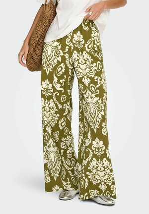 Person som har på seg olivengrønne vide bukser med stort hvitt blomsterprint, hvit topp, sølvsko og bærer en vevd brun veske.