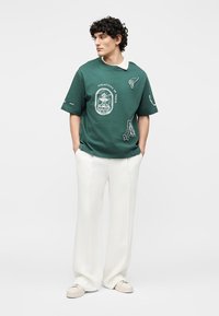 Νέος άνδρας με πράσινο γραφικό T-shirt, λευκό παντελόνι φαρδύ και μπεζ αθλητικά παπούτσια, στέκεται με τα χέρια στις τσέπες μπροστά σε απλό φόντο.