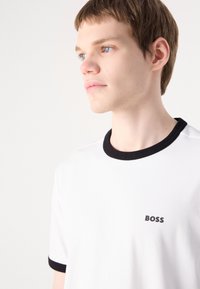 Bílý žebrovaný tričko s černým kulatým výstřihem a akcenty na rukávech, s malým černým logem "BOSS" na levé hrudi.