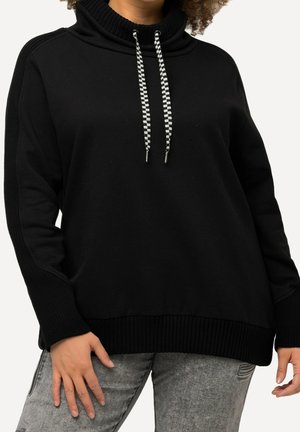 Henkilö, jolla on löysä musta huppari, jossa on ruudulliset kiristysnyörit, ja harmaat haalistuneet farkut, joissa on pieni revennyt kohta reisiosassa.