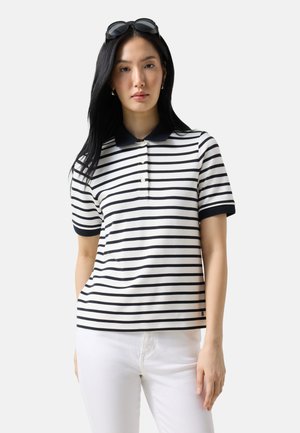 Kvinde med langt sort hår iført sorte solbriller på hovedet, en hvid og sortstribet polo-shirt og hvide bukser mod hvid baggrund.