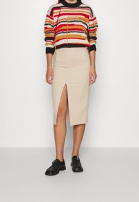 Pull en tricot multicolore avec des rayures horizontales, associé à une jupe crayon beige avec une fente sur le devant. Chaussures à lacets noires.