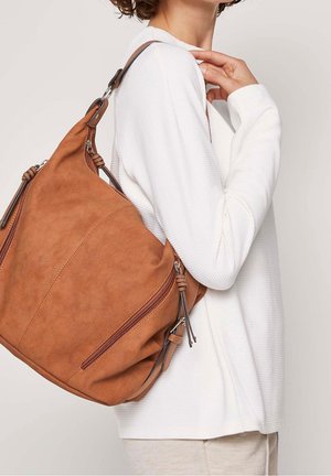 Kobieta w białej bluzce z długim rękawem, niosąca na plecach dużą beżową skórzaną torbę na ramię z zamkami błyskawicznymi.