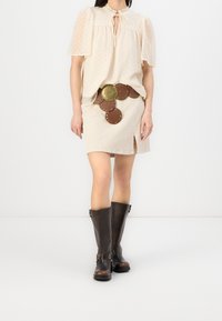 Paituro cream-colored, leher pendek dengan titik-titik tekstur, dipadukan dengan rok beige fitting yang memiliki celah samping dan ikat pinggang aksen bulat coklat. Sepatu bot coklat setinggi lutut.