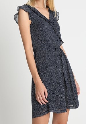 Granatowa sukienka bez rękawów w białe kropki, z marszczonym dekoltem w kształcie litery V i paskiem wiązanym w talii, noszona przez stojącą osobę.