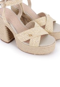 Sandalias de plataforma tejiadas en beige con correas cruzadas, un tacón grueso y una suela texturizada. Los acentos decorativos incluyen un pequeño detalle de logo.