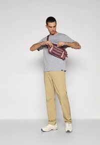 Pilka marškinėliai ir smėlio spalvos kelnės, kurias dėvi žmogus, atidarantis bordo spalvos juosmens krepšį su rožiniais akcentais ir Patagonia logotipu.