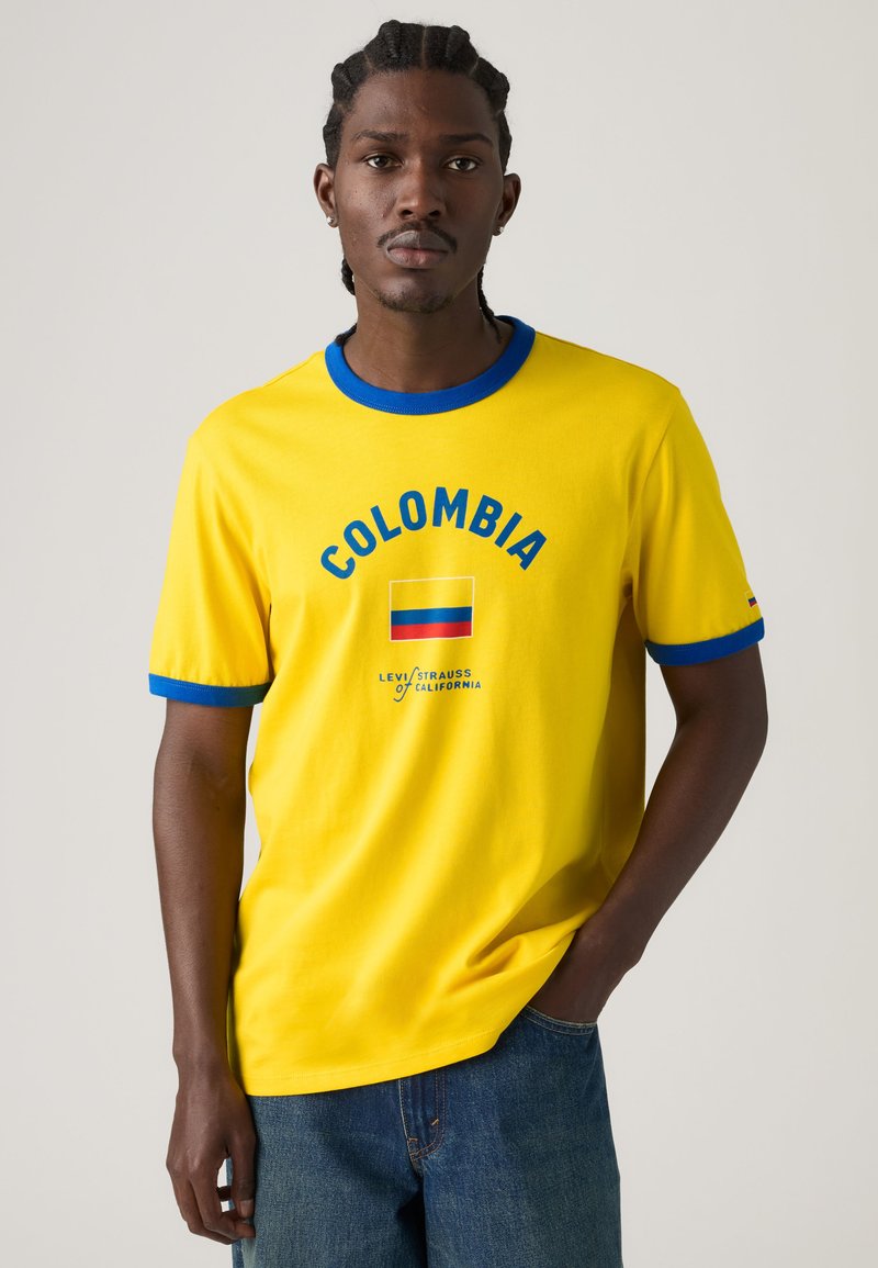 Jeune homme aux cheveux tressés portant un t-shirt jaune de la Colombie avec des bordures bleues et un jean en denim sur un fond uni.