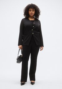 Fekete blézer arany gombokkal, hozzáillő fekete nadrággal. A modell egy kis fekete kézitáskát tart, és hegyes orrú cipőt visel.