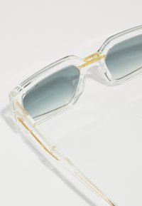 Ochelari de soare supradimensionați, cu lentile gri gradient. Accente metalice aurii la balama, ramă transparentă texturată și brațe subțiri transparente.