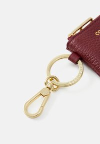 Burgundy textured leather keychain holder with a gold-colored metal clip and ring. Features embossed logo on the side, smooth edges.