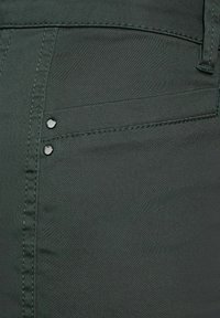 Mörkgröna byxor med en slät, strukturerad tyg och synlig sömnad samt två dekorativa svarta knappar på frontfickan.