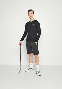 Crna dugih rukava majica, sivi šortsevi, drži golf palicu. Sadrži logotip kao akcent. Uparena s bijelim čarapama i sportskim cipelama s plavim detaljima.