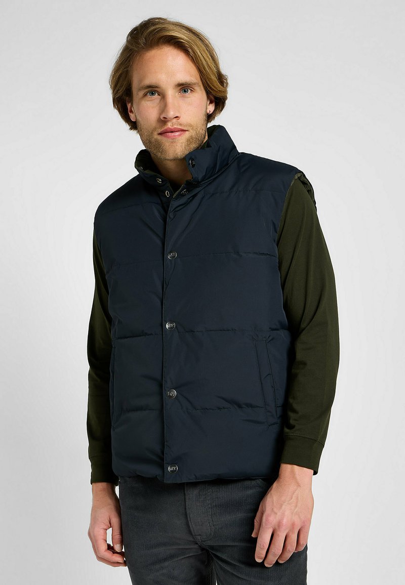 Gilet imbottito blu navy con colletto alto, chiusura con bottoni e tasche laterali, indossato sopra una camicia a maniche lunghe verde scuro.