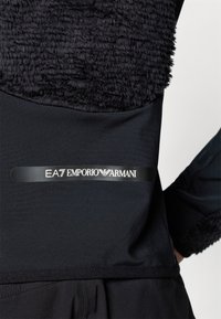 Must mustarüü, millel on pehme tekstuurne fliisiosa ja sile alumine osa. Sisaldab logoriba, kus on tekst "EA7 EMPORIO ARMANI".